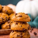 A stack of three pumpkin chocolate chip cookies on a table with a plate of more cookies in the background.