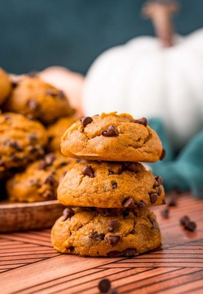 A stack of three pumpkin chocolate chip cookies on a table with a plate of more cookies in the background.