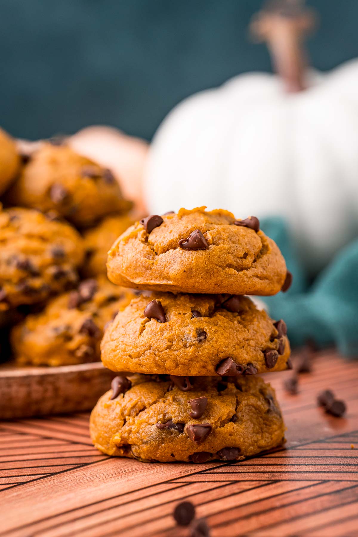 A stack of three pumpkin chocolate chip cookies on a table with a plate of more cookies in the background.