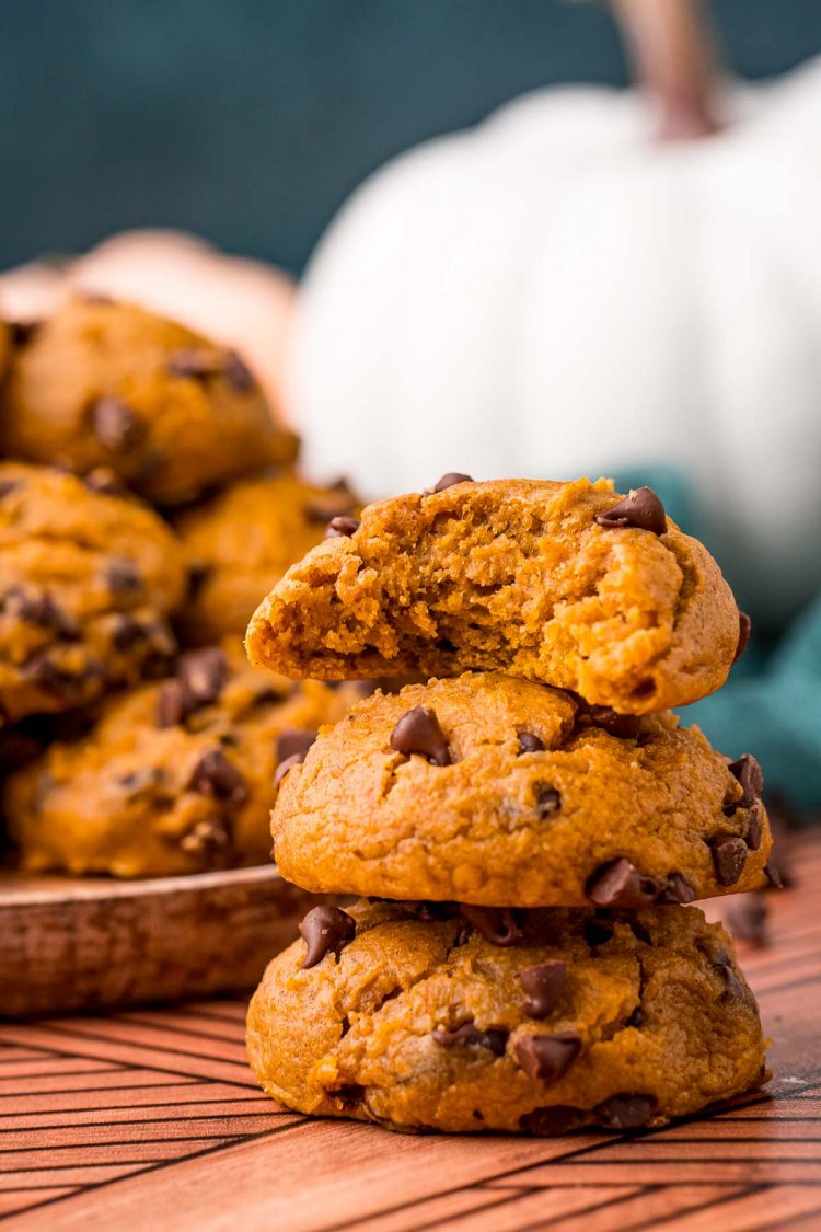 A stack of pumpkin chocolate chip cookies on a table with a bite taken out of the top on. A plate with more cookies is in the background.