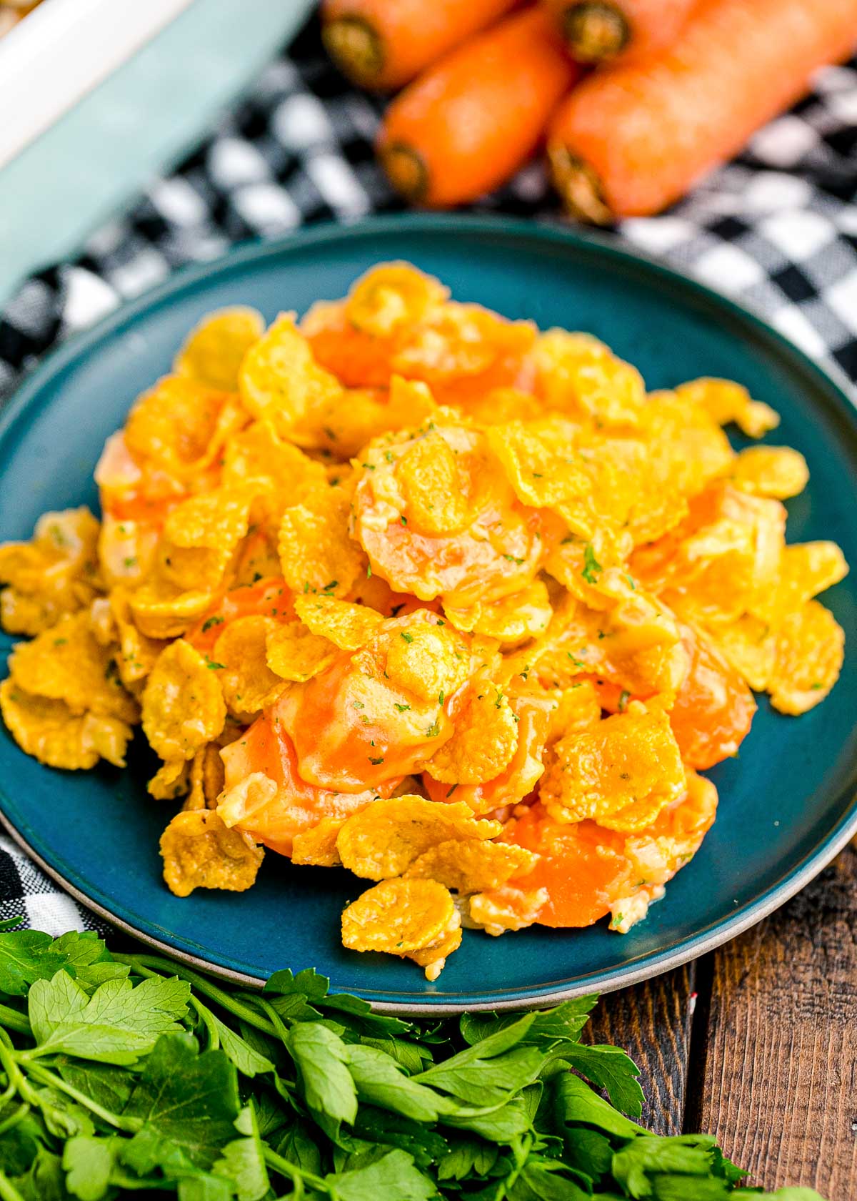 Close up photo of carrot casserole on a blue plate on a black and white checkered napkin with carrots and parsley on the side of the plate.