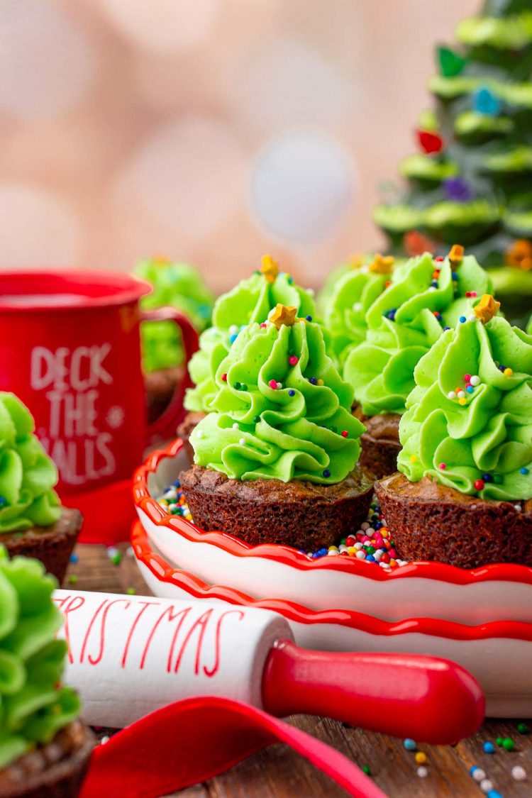 Close up photo of christmas tree brownie bites on red trimmed plates with a mug and ceramic christmas tree in the background.