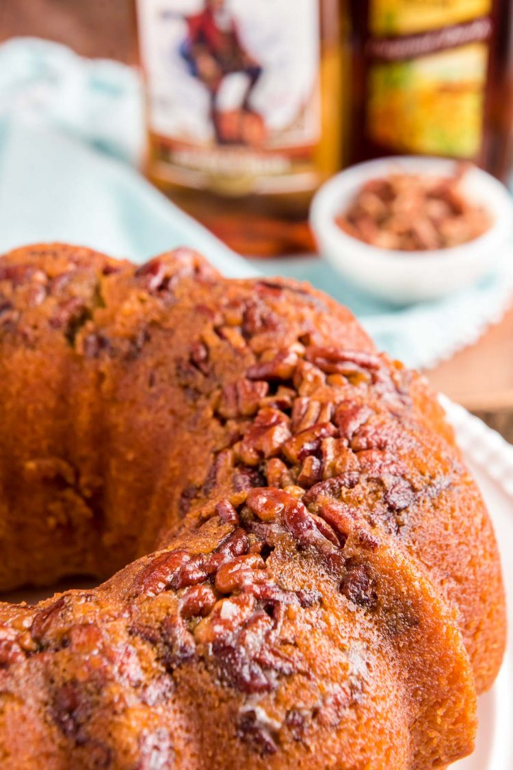 Close up photo of a rum cake on a white cake stand with bottles of rum in the background.