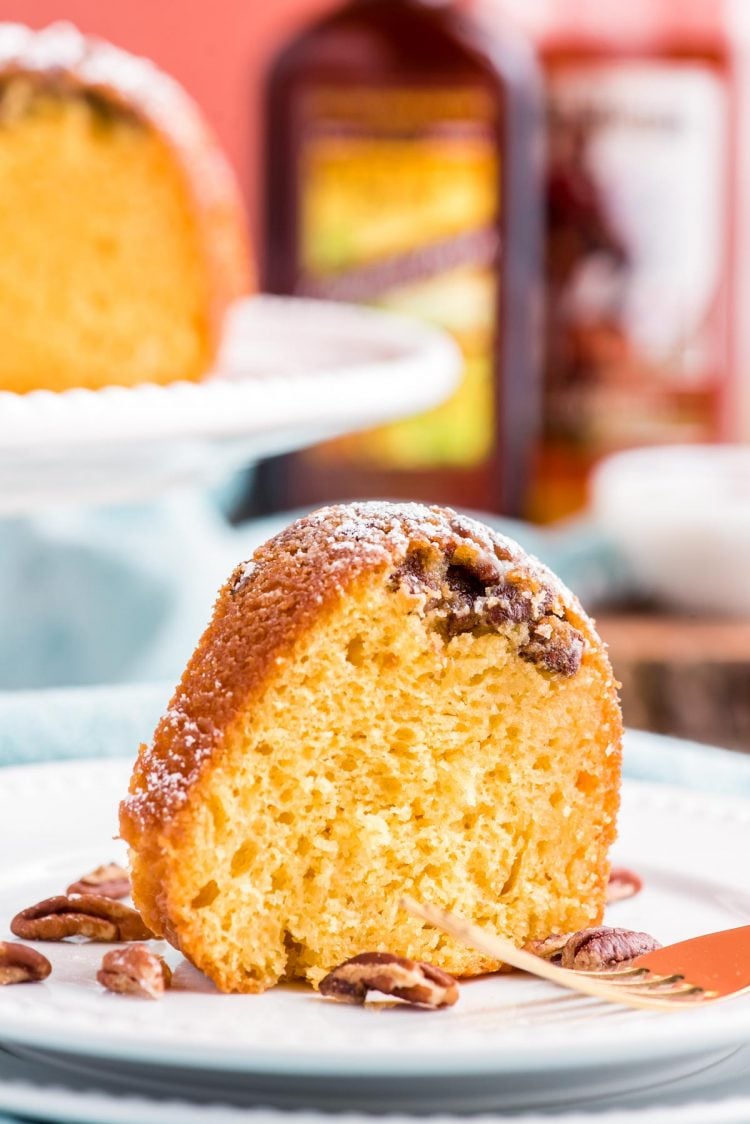 A slice of rum cake on a white plate with the rest of the cake on a white cake stand behind it. Bottles of rum are in the background.