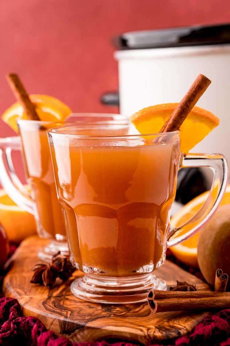 Straight on photo of two mugs of mulled cider on a wooden board with a crockpot in the background.