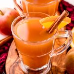 Close up photo of two mugs of mulled cider on a wooden cutting board garnished with a cinnamon stick and orange slice.