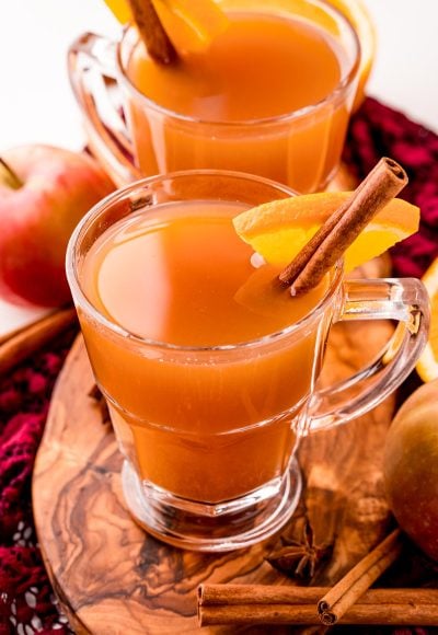 Close up photo of two mugs of mulled cider on a wooden cutting board garnished with a cinnamon stick and orange slice.