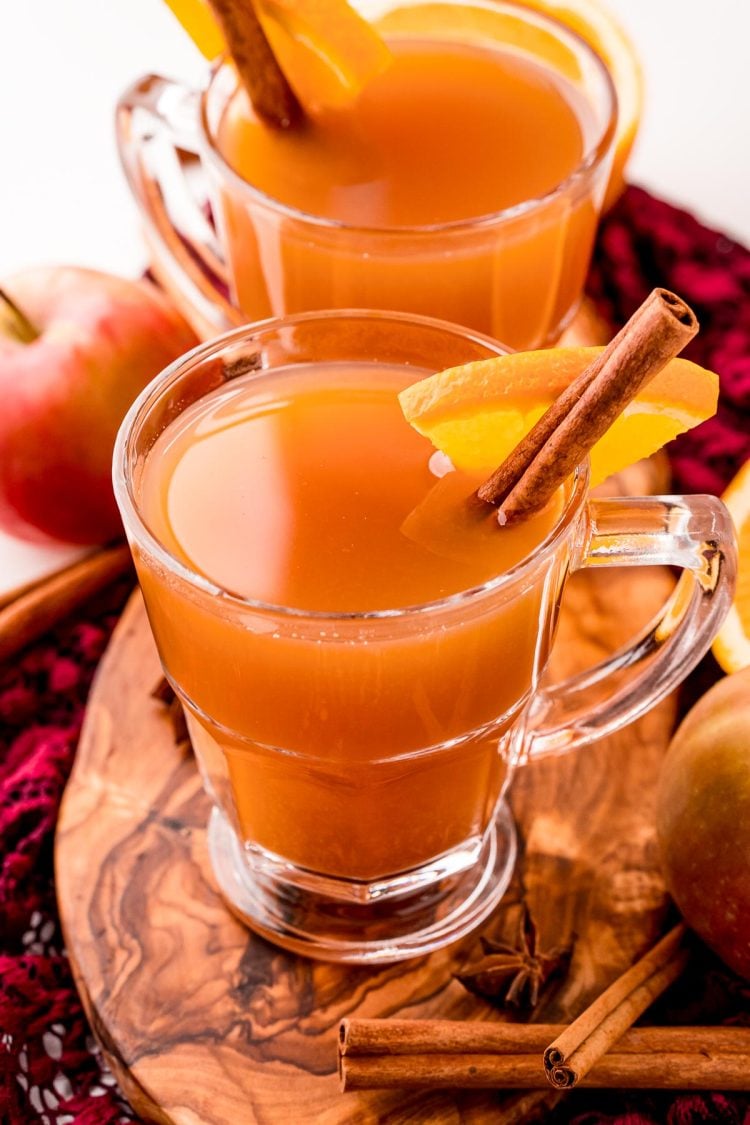 Close up photo of two mugs of mulled cider on a wooden cutting board garnished with a cinnamon stick and orange slice.