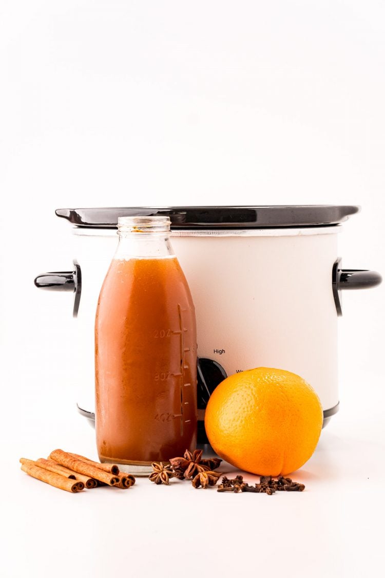 Ingredients to make mulled apple cider in front of a white crockpot on a white counter.