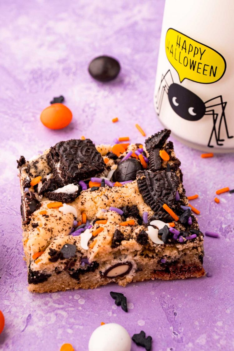 Close up photo of a slice of Halloween cookies bars on a purple surface with M&Ms and sprinkles scattered around and a small glass of milk to the side.