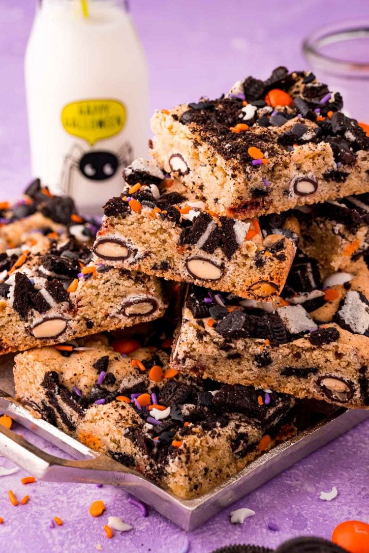 Halloween cookie bars stacked on a silver tray on a purple surface with a glass of milk in the background.