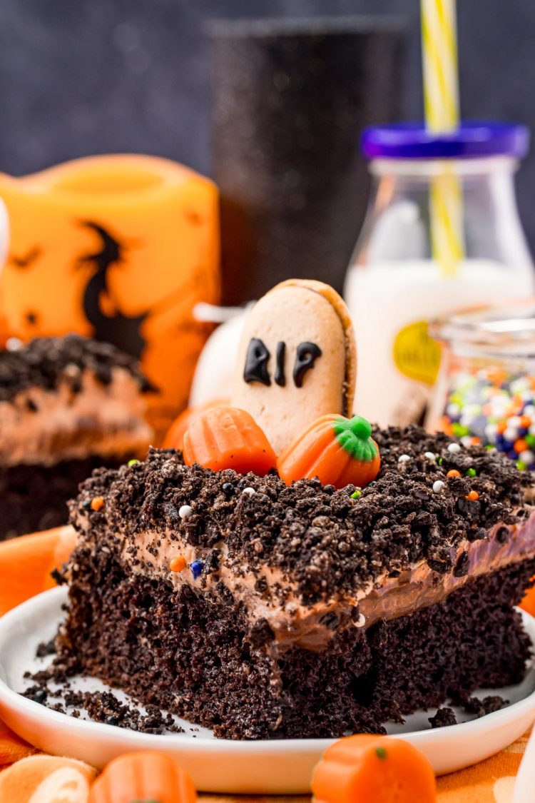 A slice of Halloween cake on a white plate on an orange napkin with halloween decorations and a bottle of milk in the background.