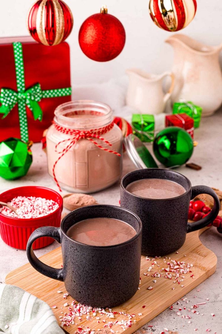 Two black mugs filed with hot chocolate one a wooden cutting board with holiday decorations in the back.