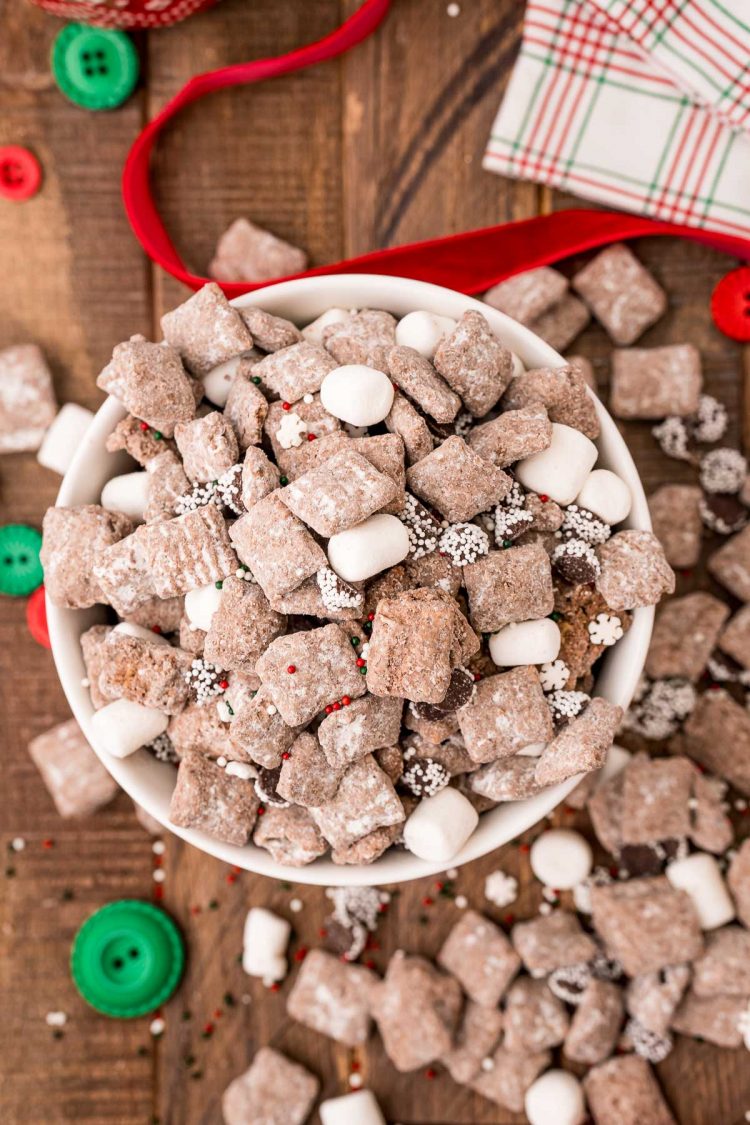 Overhead photo of a bowl of hot chocolate muddy buddies on a wooden table with holiday decorations scattered around.