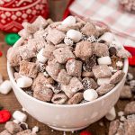 A white bowl filled with hot chocolate muddy buddies on a wooden table with muddy buddies and holiday decorations scattered around.