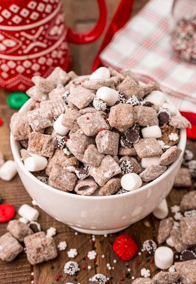 A white bowl filled with hot chocolate muddy buddies on a wooden table with muddy buddies and holiday decorations scattered around.