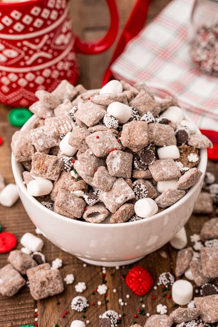 A white bowl filled with hot chocolate muddy buddies on a wooden table with muddy buddies and holiday decorations scattered around.