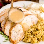 Close up photo of gravy being ladled over slices turkey breast on a white plate with sides.