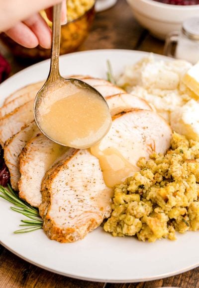 Close up photo of gravy being ladled over slices turkey breast on a white plate with sides.