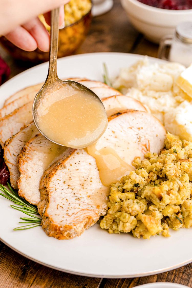 Close up photo of gravy being ladled over slices turkey breast on a white plate with sides.
