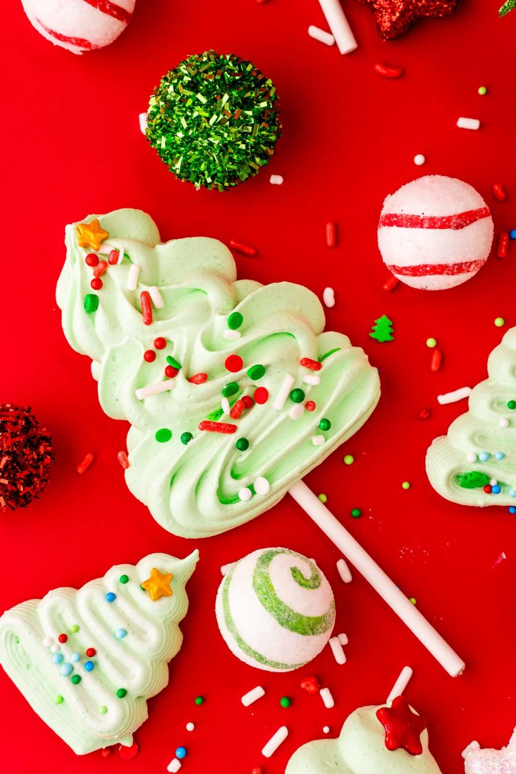Christmas Tree Mernigue cookies and cookie pops on a red surface surrounded by sprinkles and holiday decor.