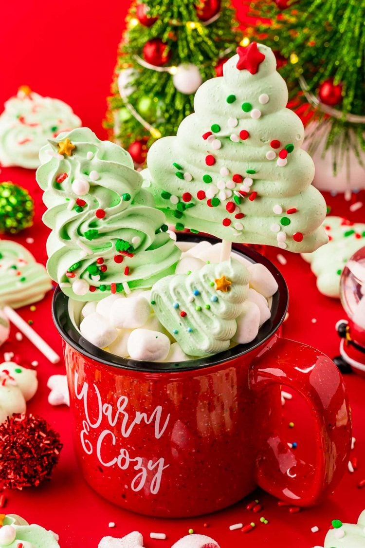 Close up photo of meringue christmas tree cookies and pops in and around a red holiday mug with decorative trees in the background.