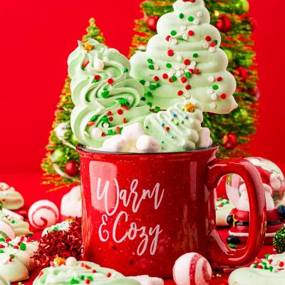 Close up photo of meringue christmas tree cookies and pops in and around a red holiday mug with decorative trees in the background.