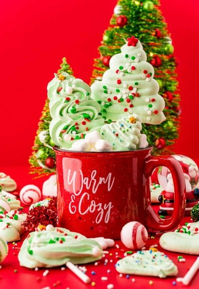 Close up photo of meringue christmas tree cookies and pops in and around a red holiday mug with decorative trees in the background.