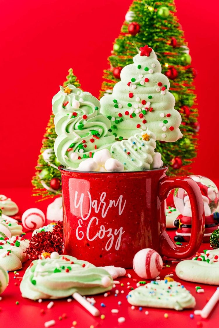Close up photo of meringue christmas tree cookies and pops in and around a red holiday mug with decorative trees in the background.