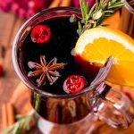 Close up photo of a glass mug filled with mulled wine garnished with rosemary and orange slice.