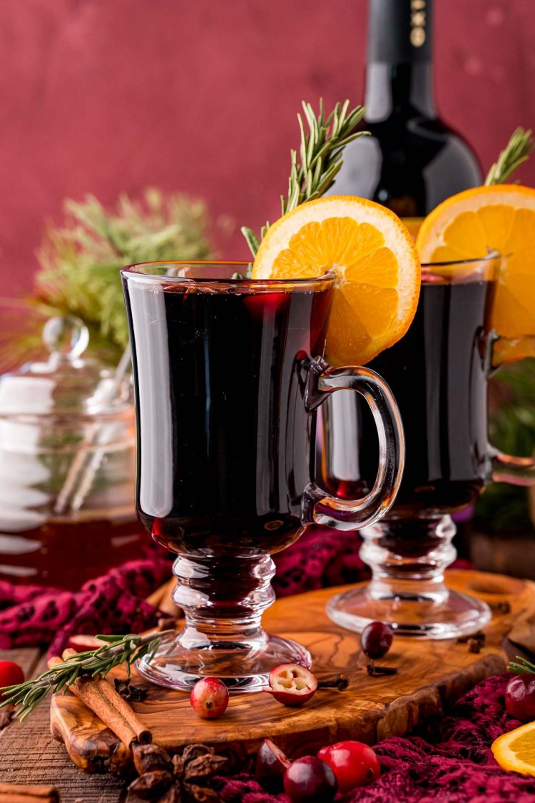 Straight on photo of two glass mugs of mulled wine on a wooden board with honey and a bottle of wine in the background.