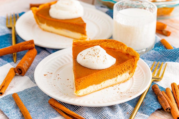 A slice of pumpkin cheesecake pie on a white plate on a blue napkin with silverware and cinnamon stick around it and another slice of pie and glass of milk in the background.