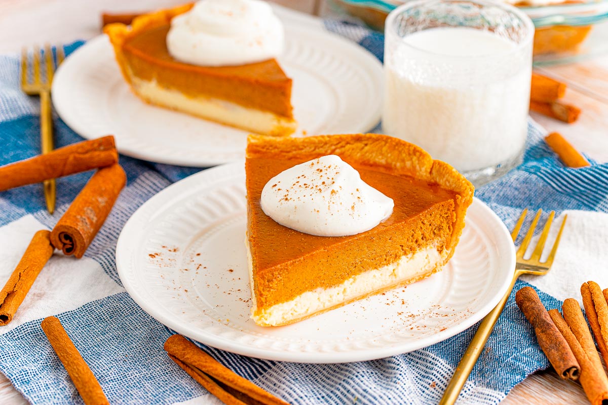 A slice of pumpkin cheesecake pie on a white plate on a blue napkin with silverware and cinnamon stick around it and another slice of pie and glass of milk in the background.