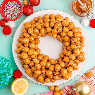 Overhead photo of struffoli on a white plate on a mint green surface with holiday decorations scattered around.