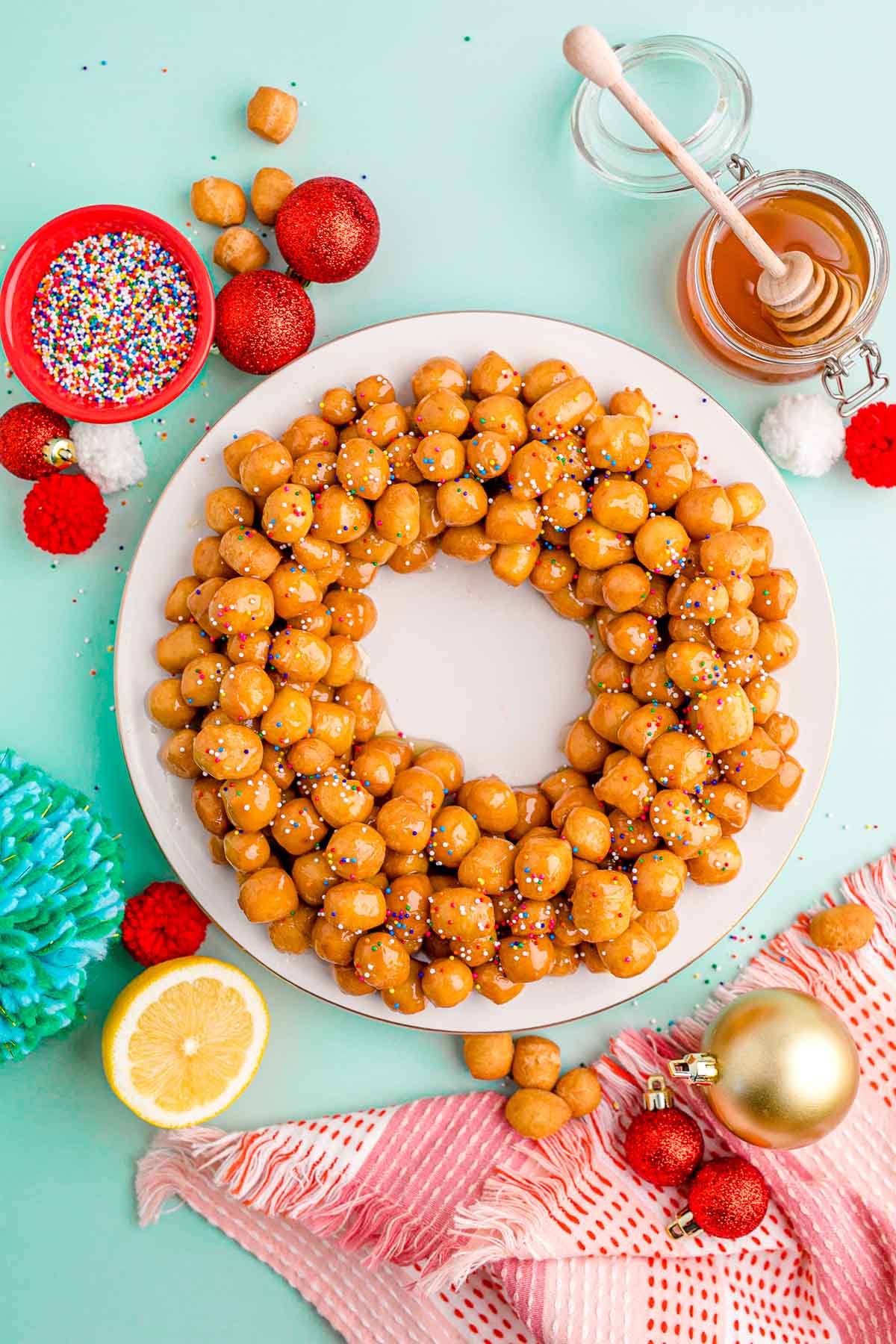 Overhead photo of struffoli on a white plate on a mint green surface with holiday decorations scattered around.