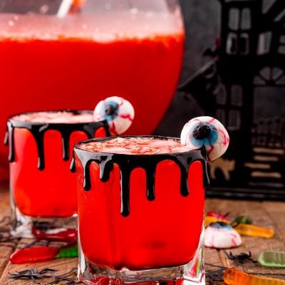 Photo of two glasses with Werewolf Halloween punch in them on a wooden table surrounded but gummy worms and eyeball gummies with a large punch bowl in the background.
