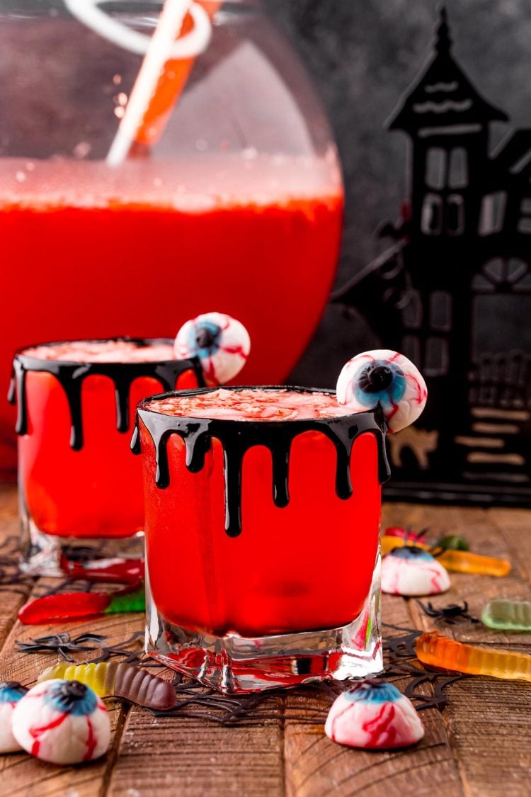 Photo of two glasses with Werewolf Halloween punch in them on a wooden table surrounded but gummy worms and eyeball gummies with a large punch bowl in the background.