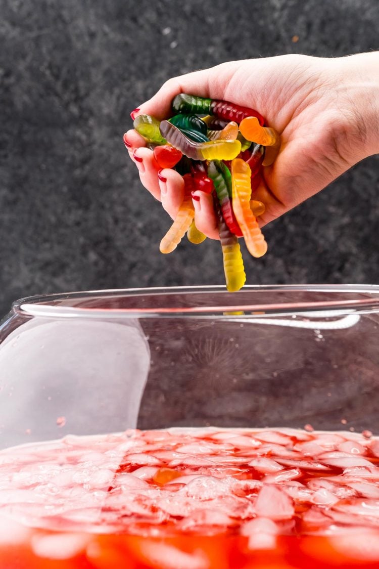 A woman's hand filled with gummy worms over a punch bowl filled with red punch ready to drop them in.