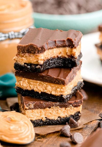 Close up photo of a stack of three buckeye brownies on a wooden table with chocolate chips and peanut butter around it.