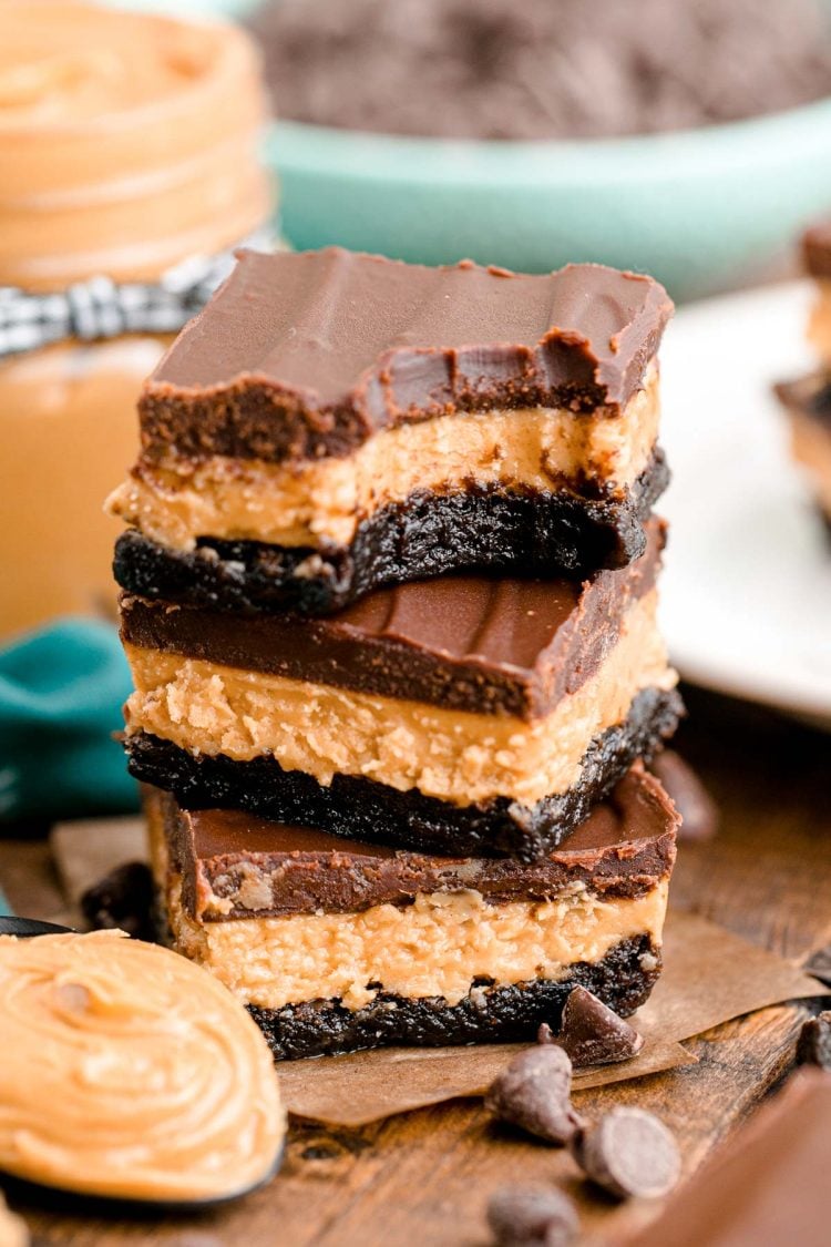 Close up photo of a stack of three buckeye brownies on a wooden table with chocolate chips and peanut butter around it.
