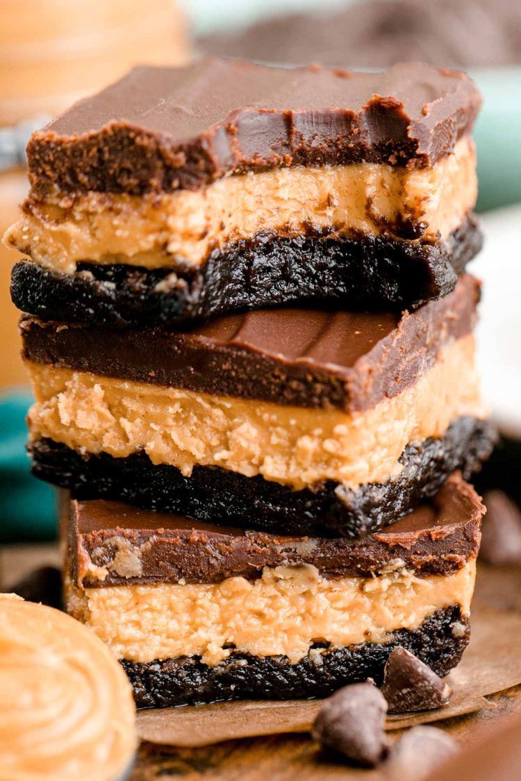 Close up photo of a stack of three buckeye brownies on a wooden table with chocolate chips and peanut butter around it.