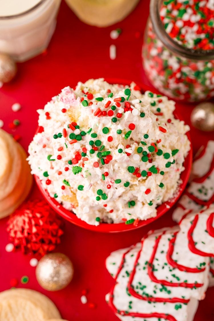 Close up photo of christmas tree cake dip in a red bowl on a red surface with holiday decorations scattered around.