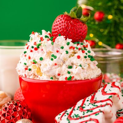 Close up photo of little debbie christmas tree dip in a red bowl on a red surface surrounded but holiday decorations with a strawberry dipped into it.