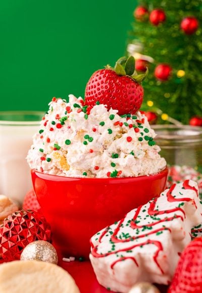 Close up photo of little debbie christmas tree dip in a red bowl on a red surface surrounded but holiday decorations with a strawberry dipped into it.