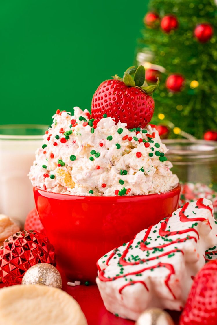 Close up photo of little debbie christmas tree dip in a red bowl on a red surface surrounded but holiday decorations with a strawberry dipped into it.