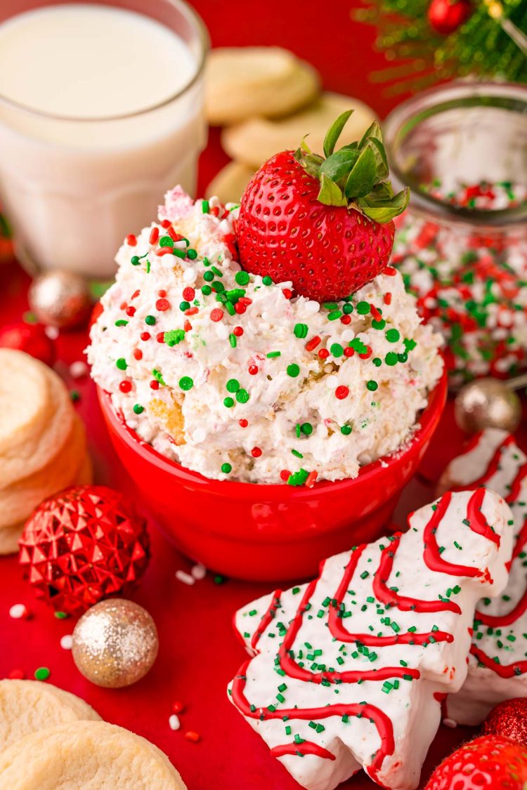 Close up photo of little debbie christmas tree dip in a red bowl on a red surface surrounded but holiday decorations with a strawberry dipped into it.