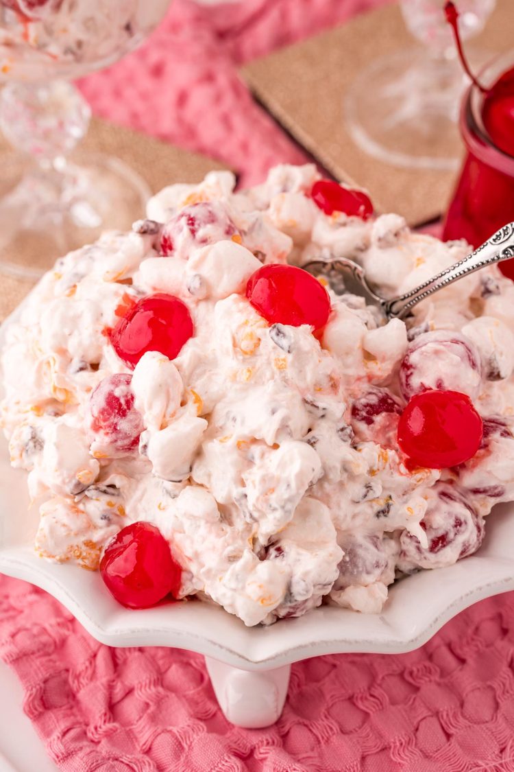 Ambrosia salad in a white serving dish with a silver serving spoon on a pink napkin.