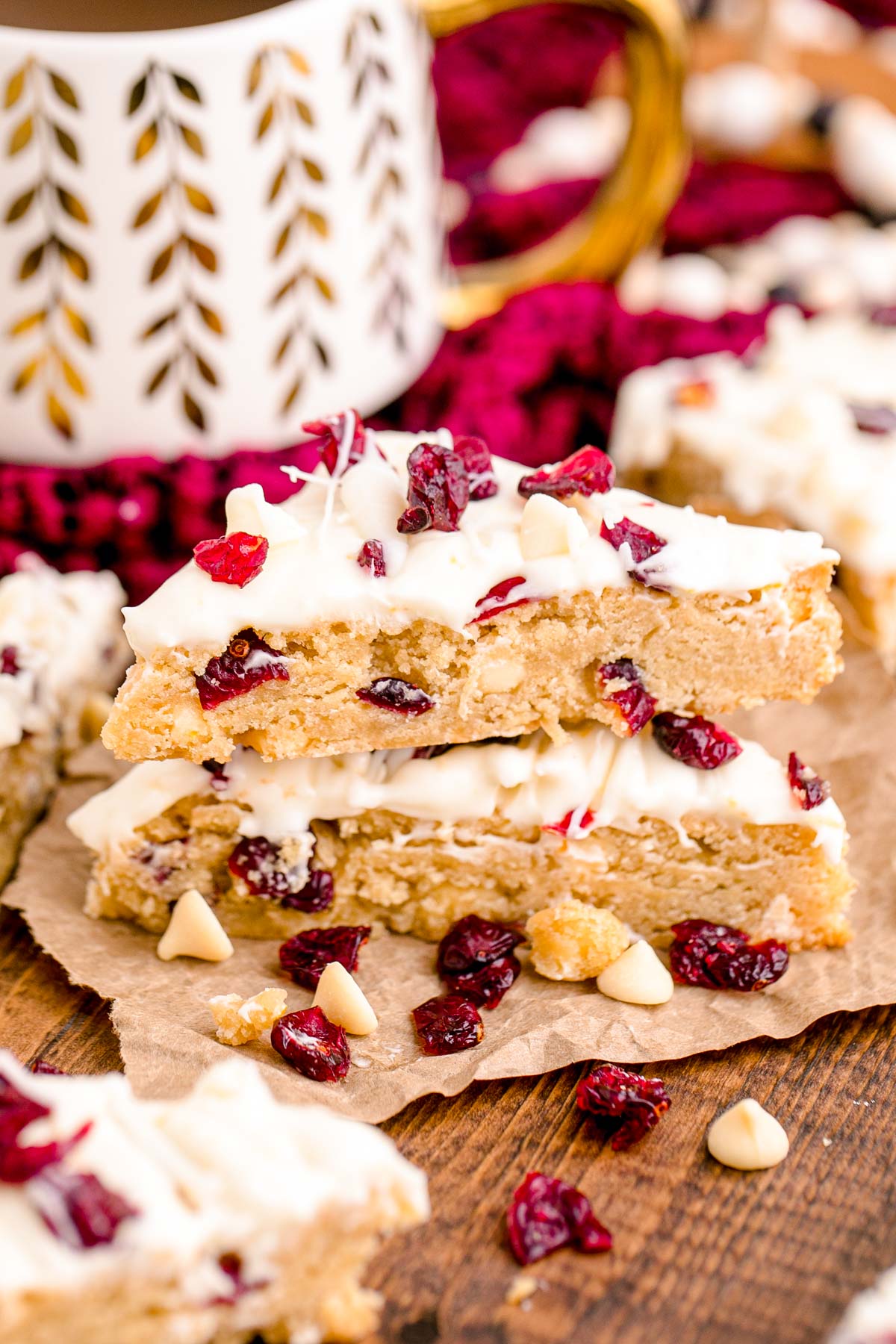 Cranberry Bliss Bars on a wooden table with cranberries and white chocolate chips scattered around with a holiday mug in the background.