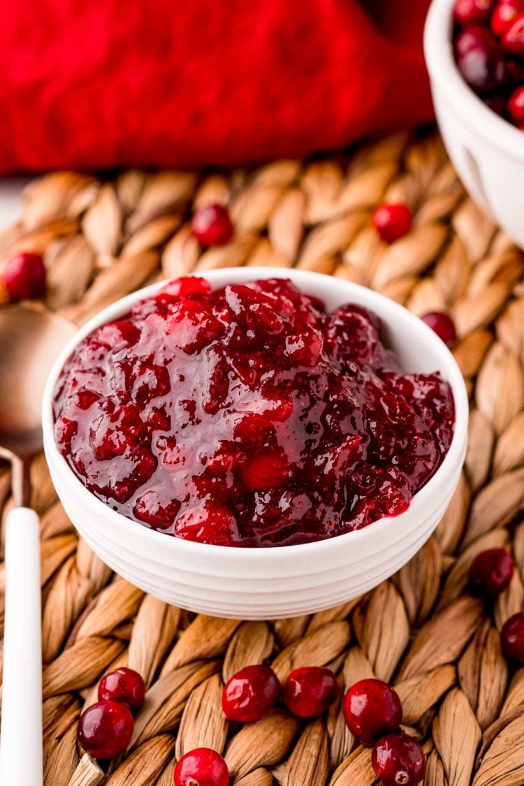 Close up photo of a what bowl filled with cranberry sauce on a whicker placemat with cranberries scattered around it.