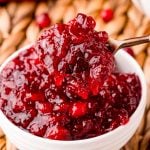 A spoon scooping cranberry sauce out of a white bowl on a wicker placemat.